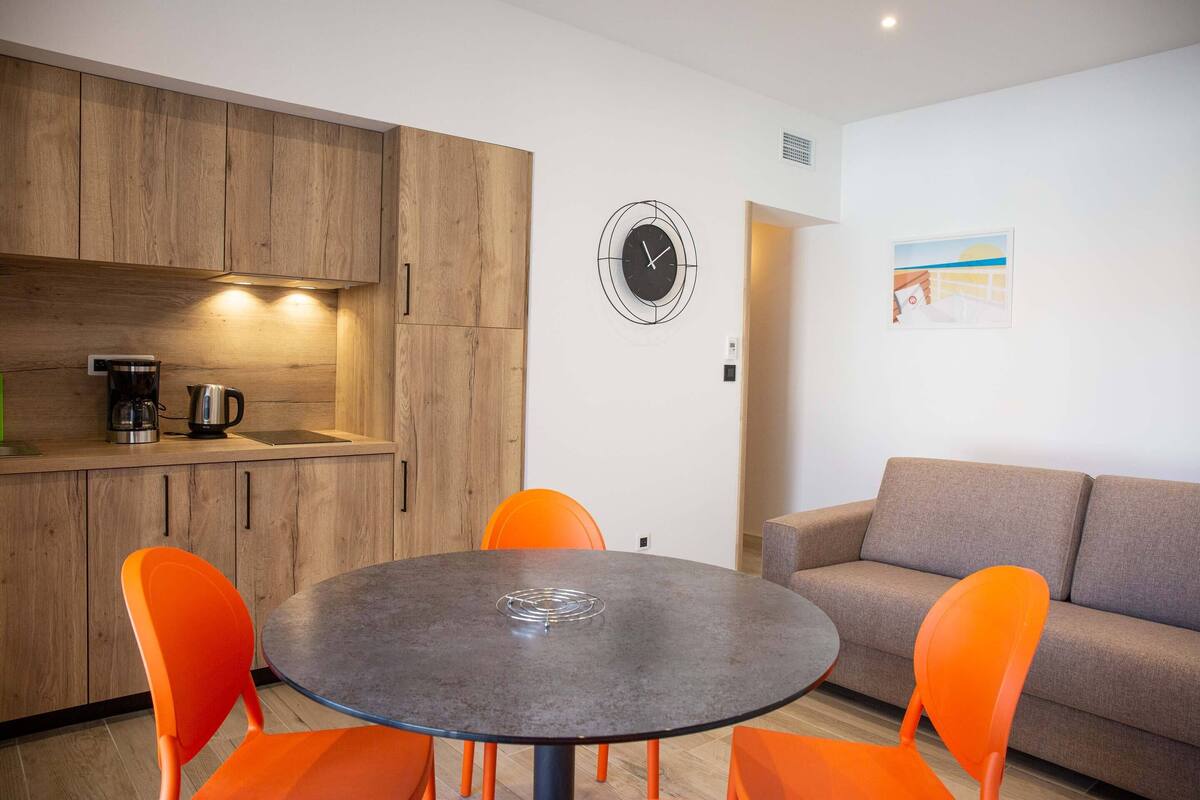 A modern kitchen with wooden cabinets, a round table with orange chairs, a clock, and a sofa in a living area.