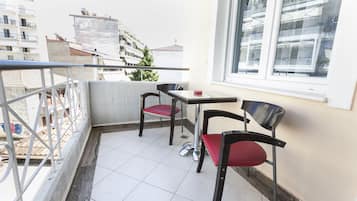 A balcony with two chairs and a table, a view of buildings, and a window with blinds.