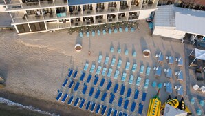 Vista aérea de un hotel frente a la playa con filas de tumbonas, un área de piscina y un edificio con varios balcones.