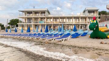 A beachfront hotel with multiple balconies, rows of blue sun loungers, and a large inflatable dinosaur decoration.