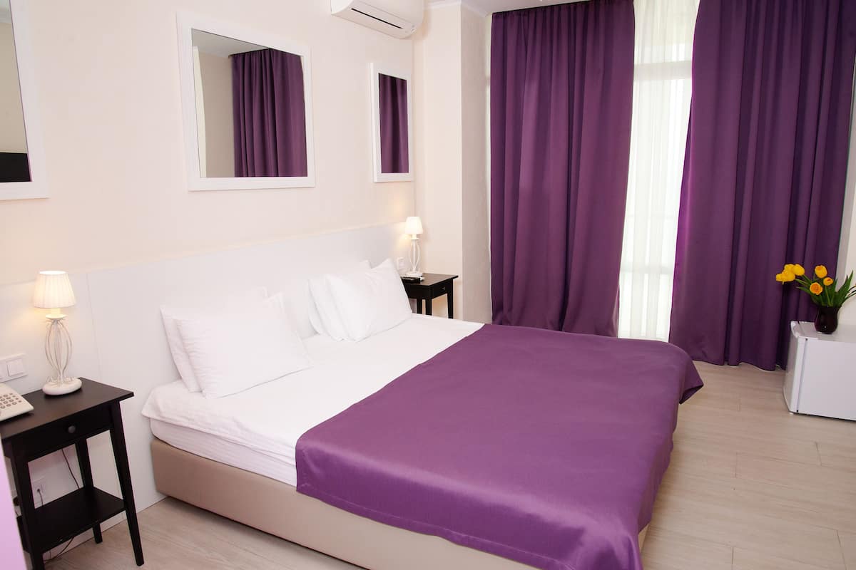 Habitación de hotel con una cama grande, una mesita de noche con lámpara, una pequeña mesa con un jarrón de flores y cortinas moradas.