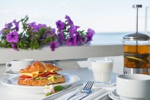 A breakfast setting with a croissant sandwich, a glass of milk, a cup of coffee, and a teapot on a table with a view of the sea and purple flowers in the background. - Design - hotel Skopeli (Odesa)