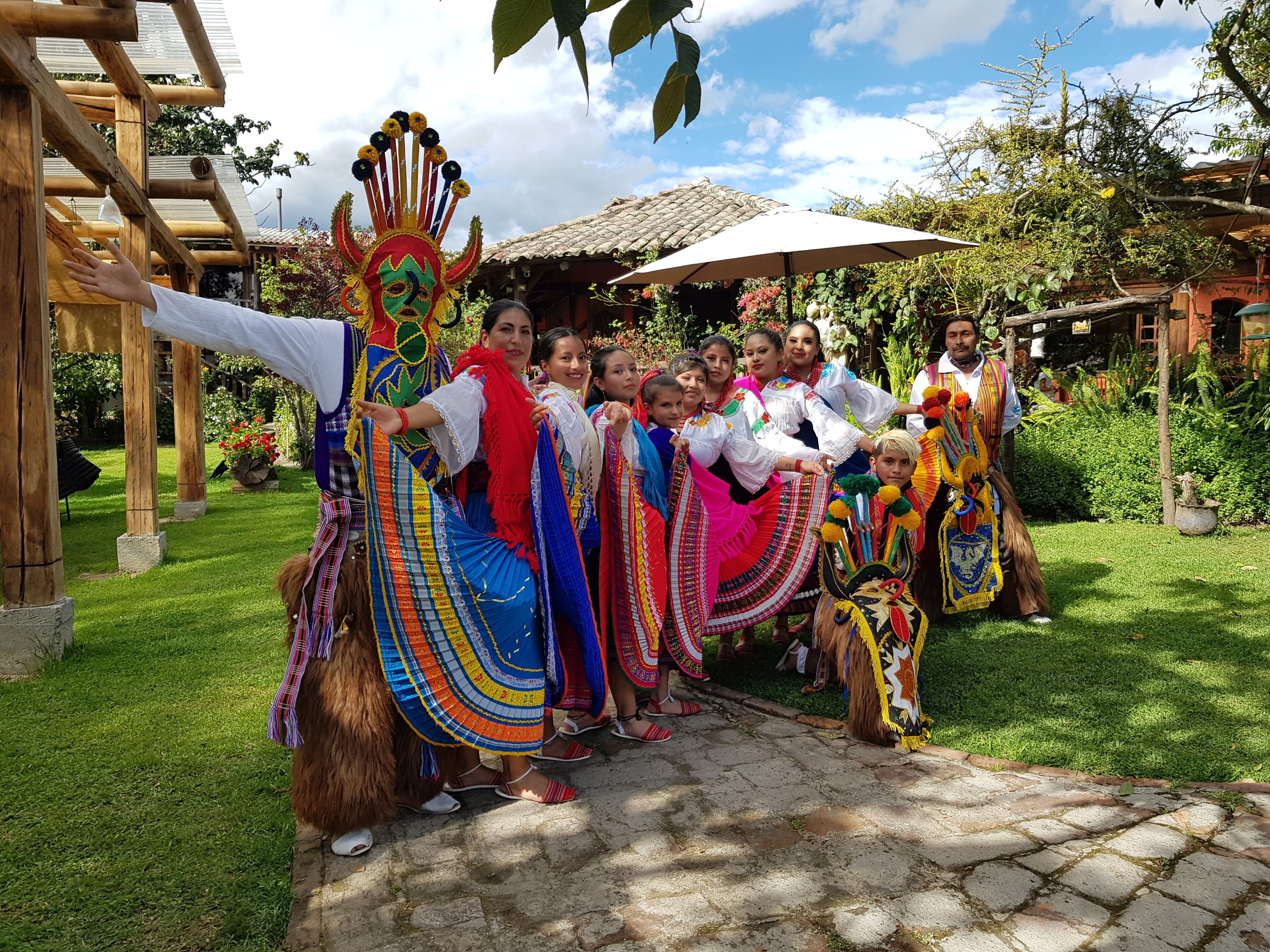 Un groupe de personnes, vêtues de costumes traditionnels aux couleurs vives, posent dans un jardin, avec une pergola en bois et une structure à toit de chaume en arrière-plan.
