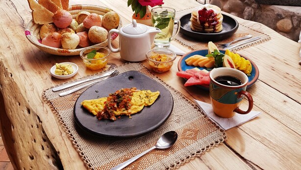 Mesa de desayuno al aire libre con pan, bollería, fruta y bebidas.