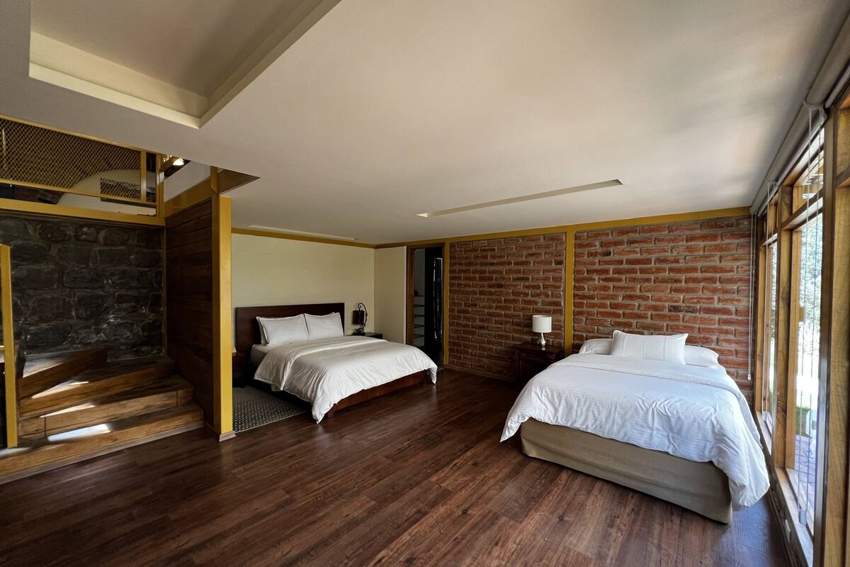 A spacious bedroom with two beds, wooden flooring, a brick accent wall, and large windows.