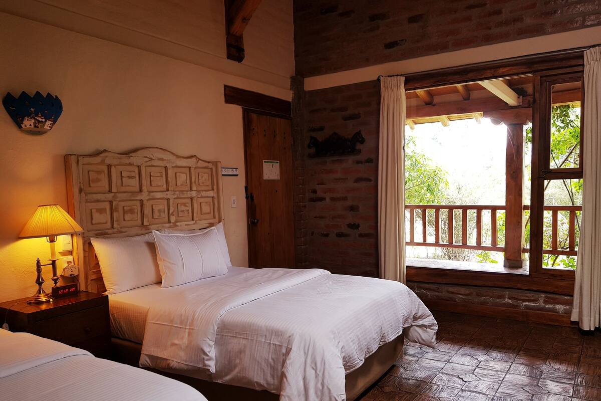 A room with two beds, a wooden headboard, a nightstand with a lamp, and a window with a view of greenery.