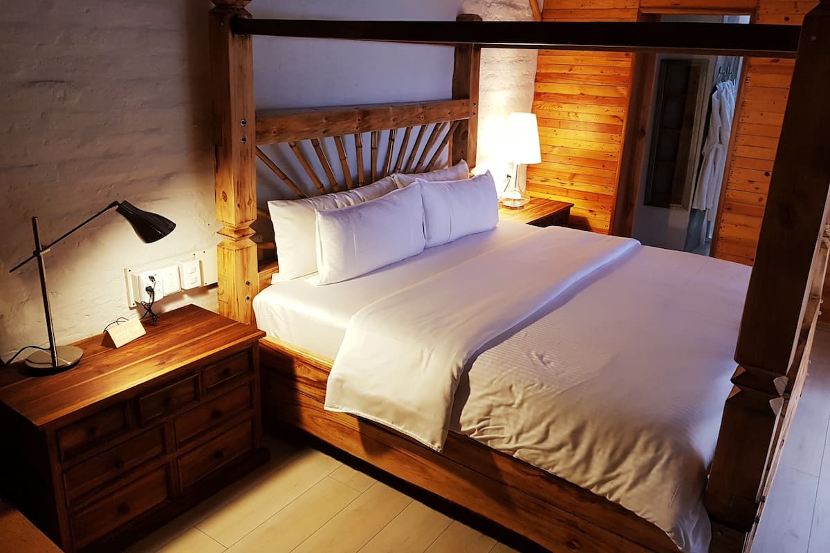 A wooden four-poster bed with white linens, a nightstand with a lamp, and a wooden dresser.