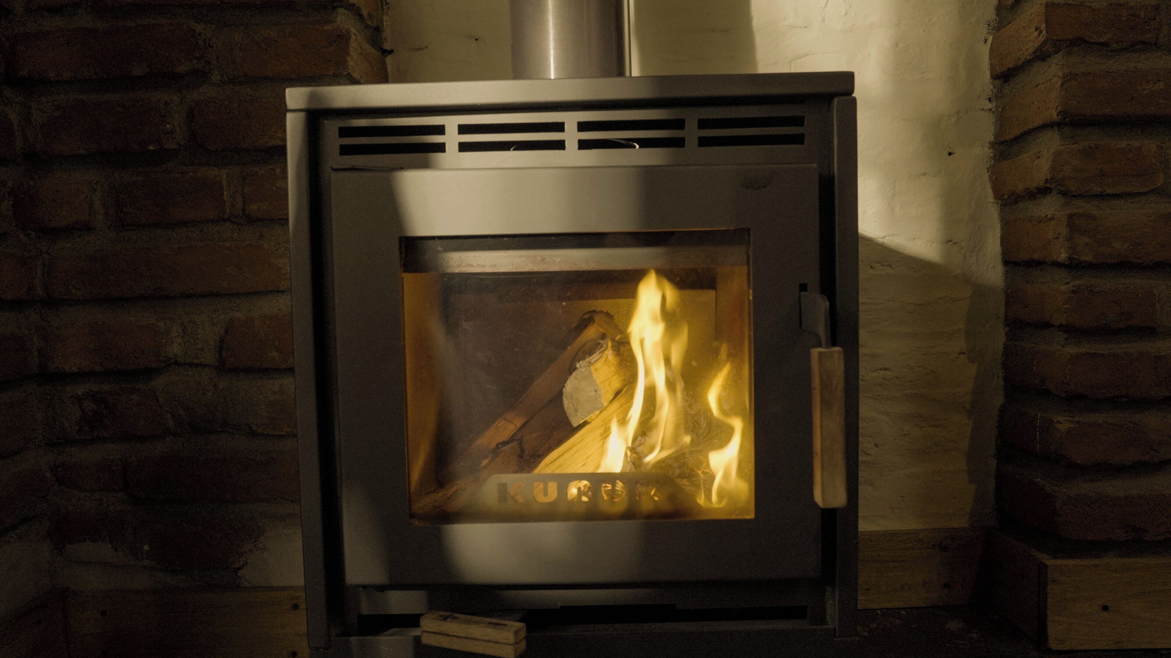 A wood-burning stove with visible logs and flames, set against a brick wall.