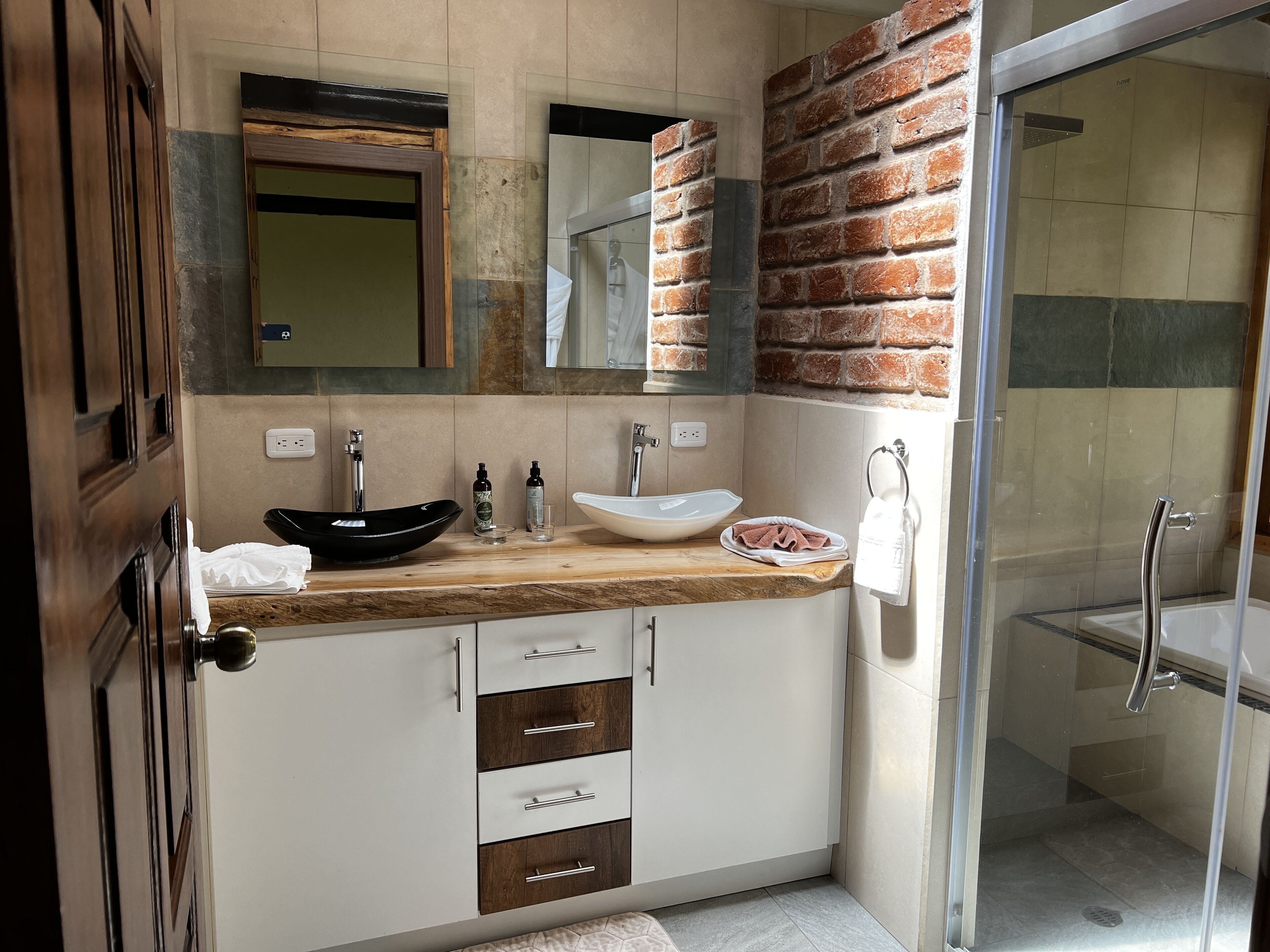 A modern bathroom with a wooden vanity, a black vessel sink, a white basin, a glass shower enclosure, and a glass door leading to a shower area.