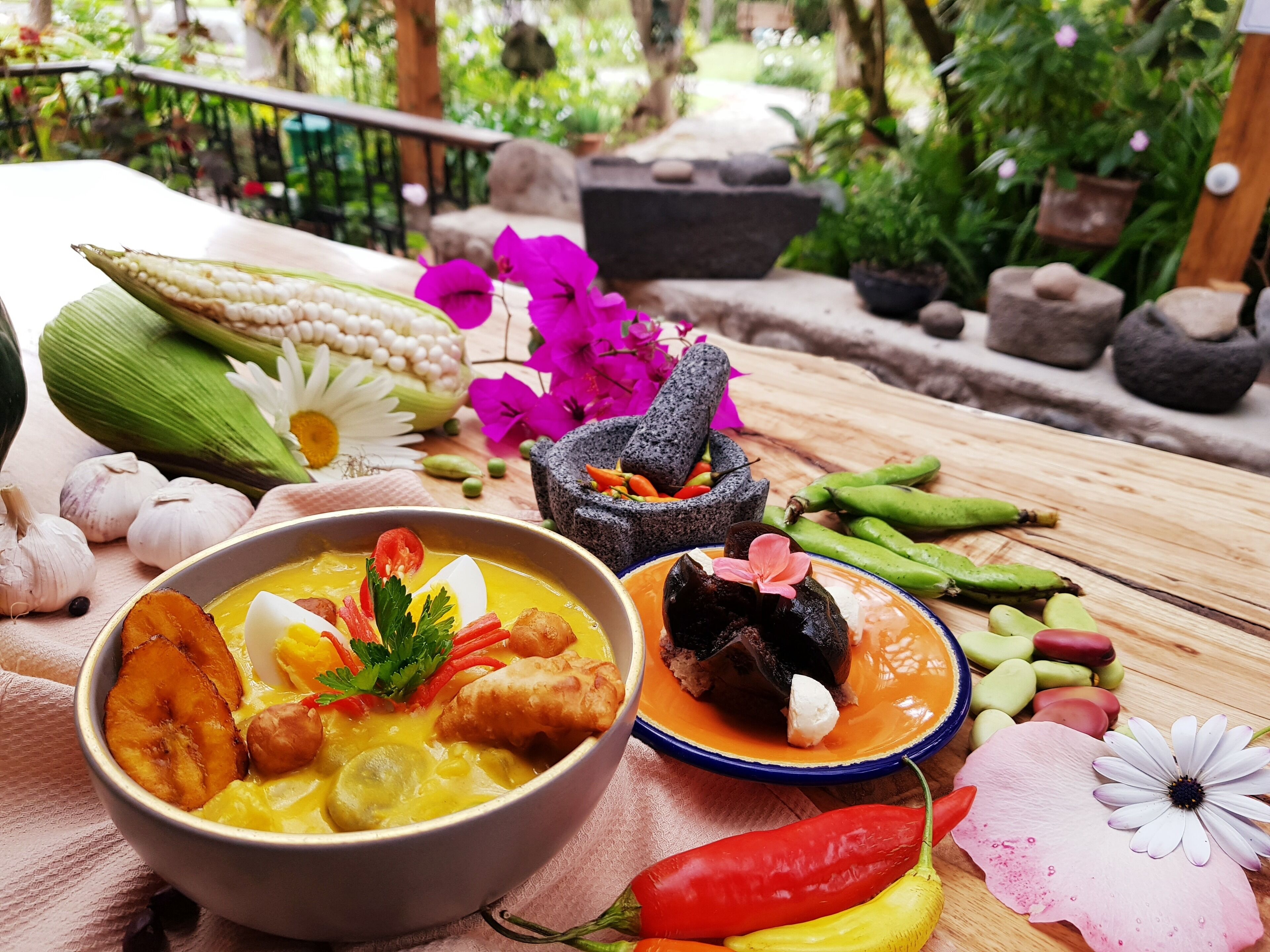 A table with a bowl of soup, a plate of food, and various vegetables and spices.