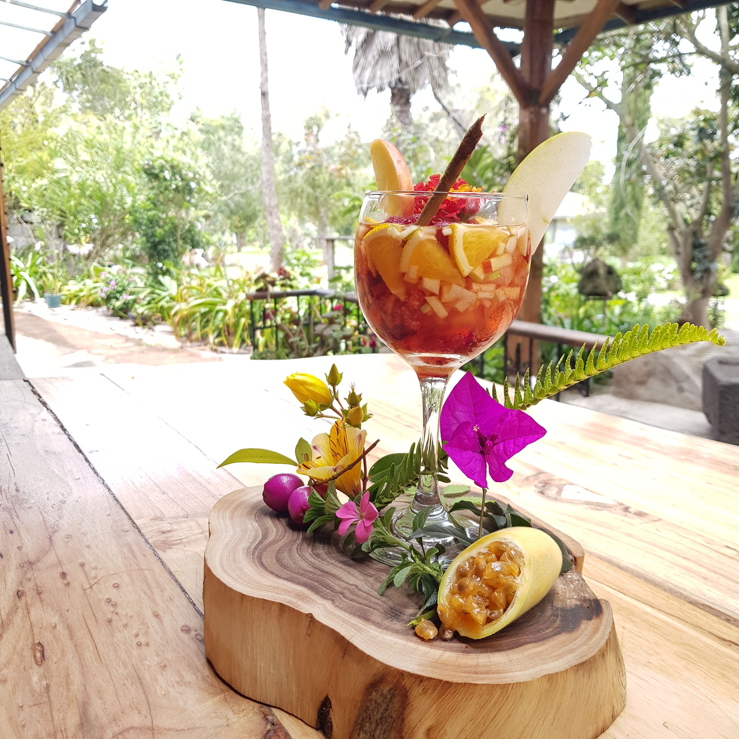 A tropical drink on a wooden board with a glass, surrounded by flowers and a half-cut fruit.