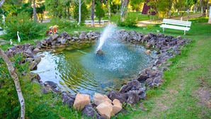 A pond with a fountain, surrounded by rocks and greenery, with a bench and a statue in the background.