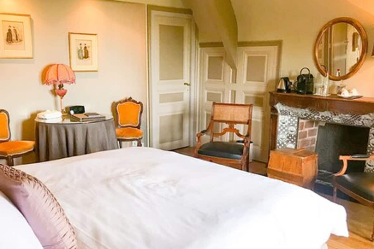 Une chambre à coucher avec un grand lit, une cheminée, deux fauteuils, une table et des tableaux encadrés accrochés au mur.