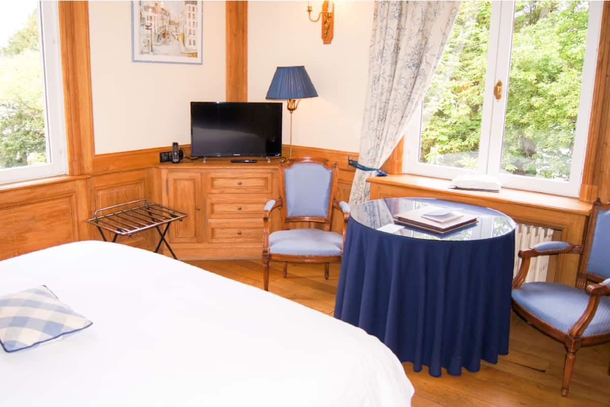 Une chambre d’hôtel avec un lit, un bureau avec une télévision, une chaise, une petite table ronde recouverte d’une nappe bleue et une fenêtre avec des rideaux.
