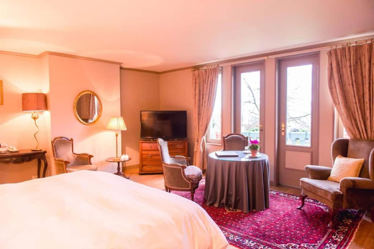 Une chambre d’hôtel avec un grand lit, une télévision, une table ronde avec une chaise, et une vue sur l’extérieur grâce à de grandes fenêtres.