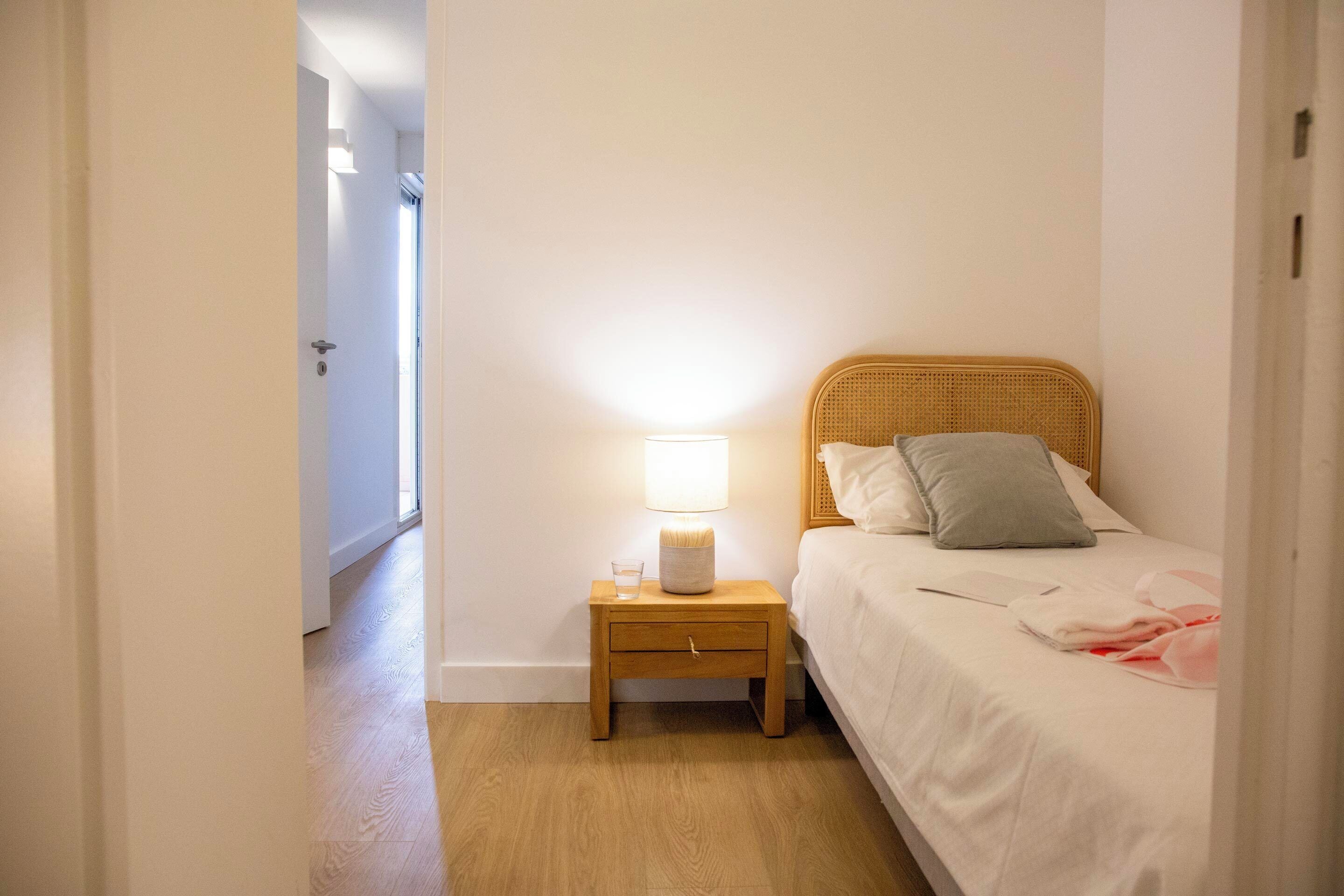A bedroom with a wooden nightstand, a bedside lamp, a bed with white bedding and a grey pillow, and a glass of water on the nightstand.
