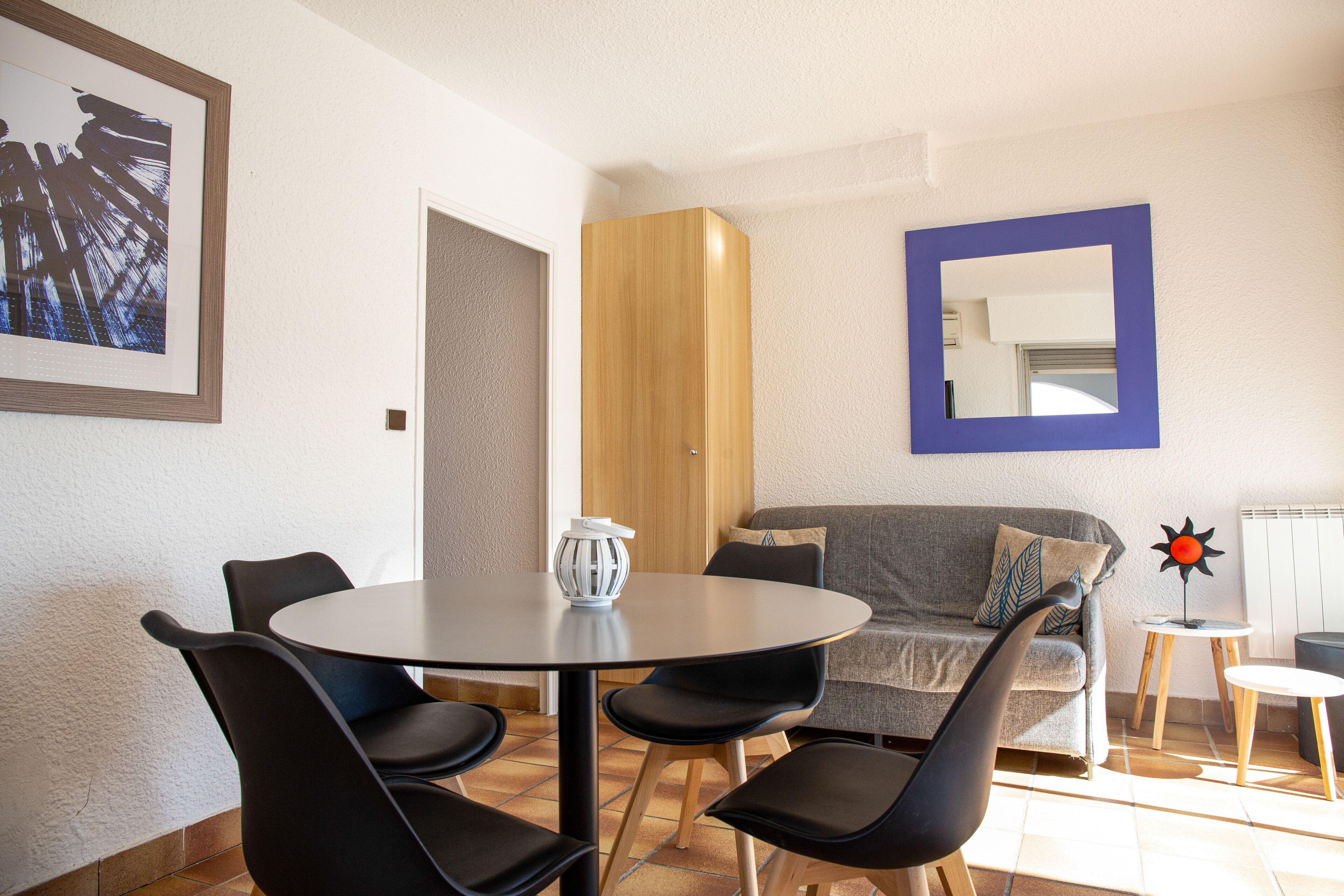 A dining area with a round table and four chairs, a sofa, a wooden cabinet, and a blue-framed mirror.