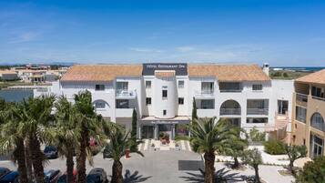 A hotel with a tiled roof, a central entrance, and a parking area with cars and palm trees.
