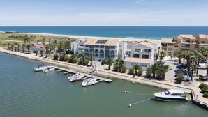 Un port de plaisance où sont amarrés plusieurs bateaux, un bâtiment à l’architecture moderne et, au loin, une plage.