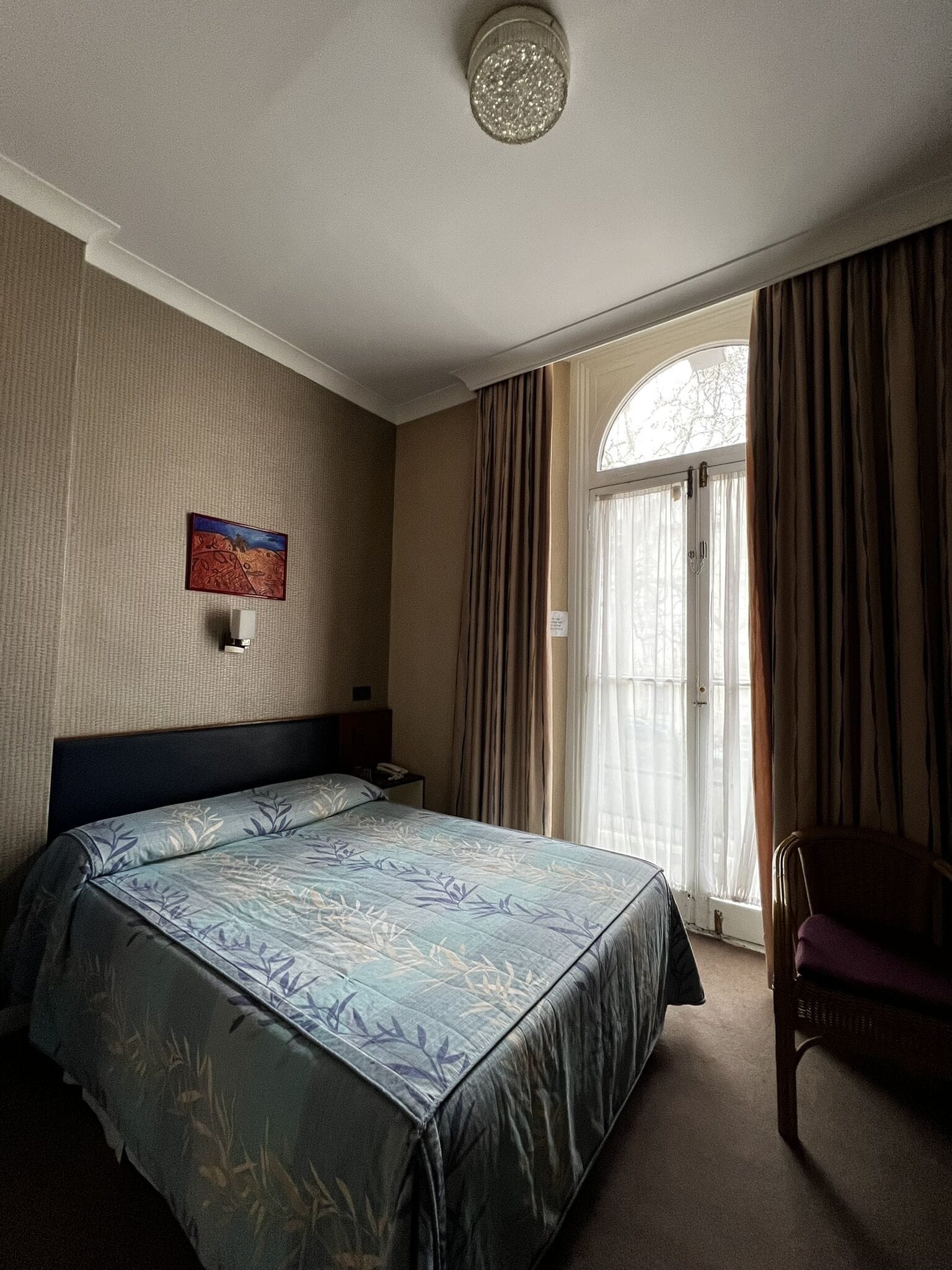 Ein Hotelzimmer mit Bett, Nachttisch, Stuhl, einem Fenster mit Vorhängen und einer Wandleuchte.