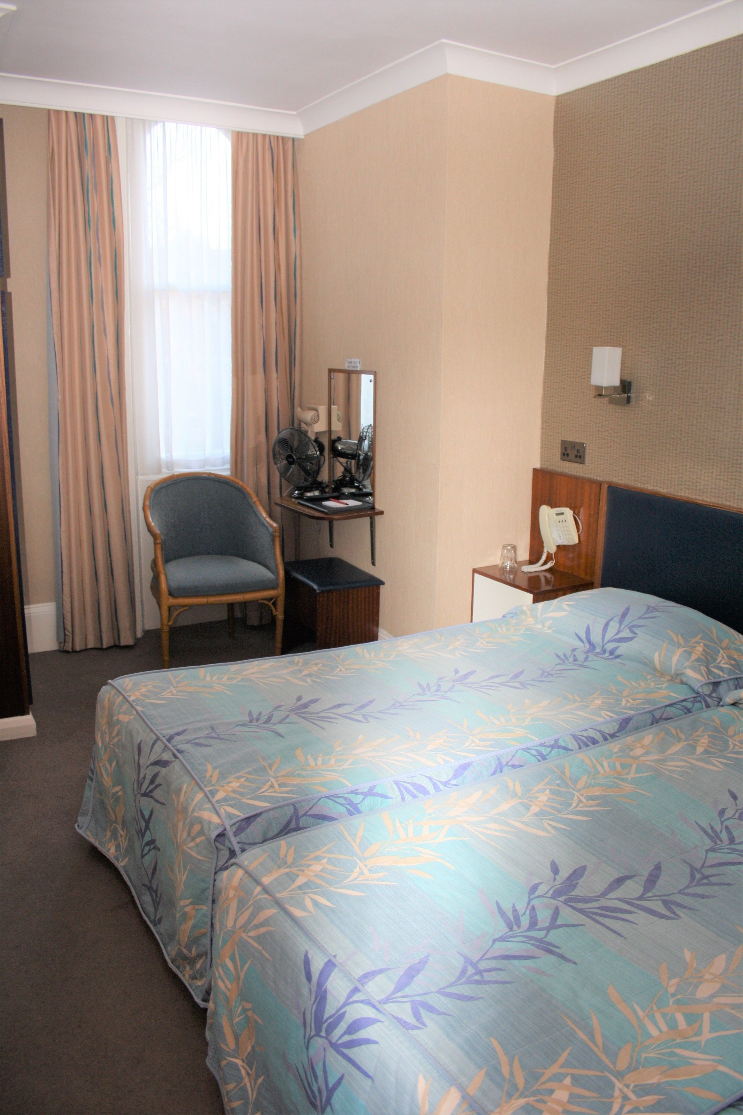A hotel room with a bed, a chair, a desk with a fan, a telephone, and a window with curtains.