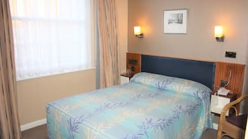 A hotel room with a bed, a nightstand, a chair, a window with curtains, and wall-mounted lights.
