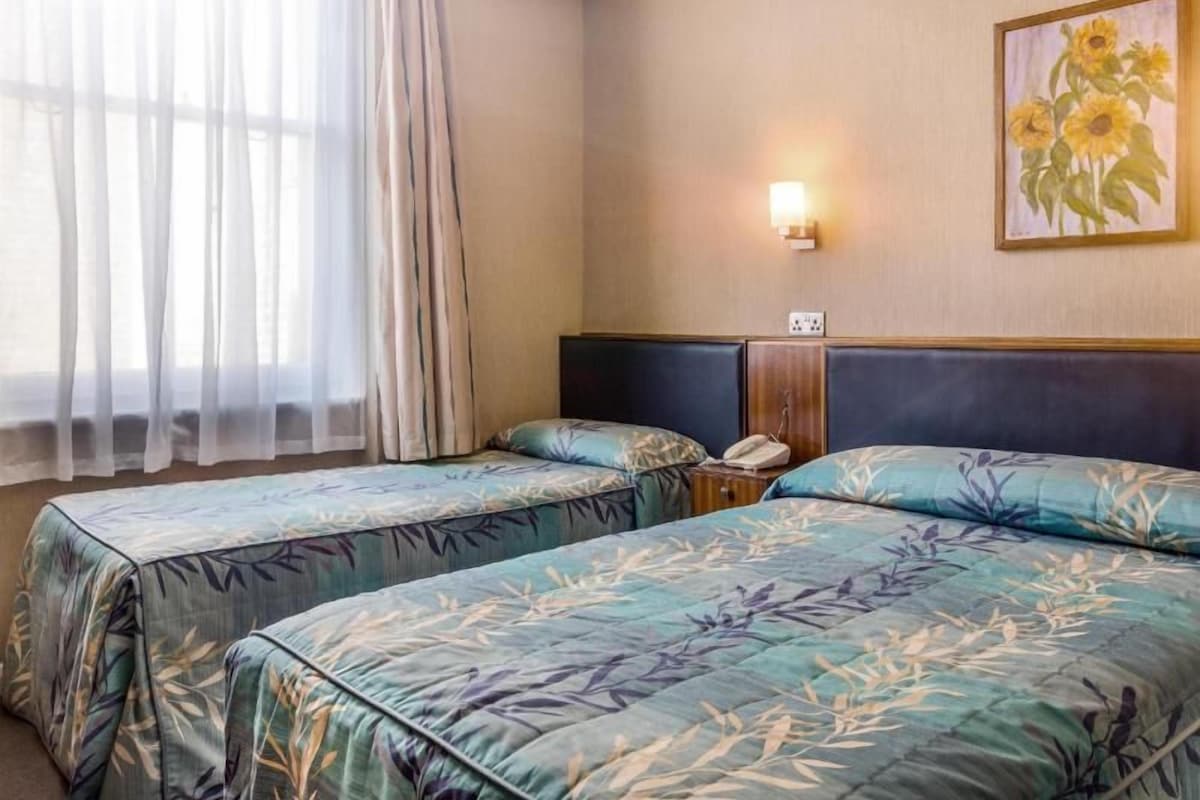 A hotel room with two beds, a nightstand, a lamp, a painting of sunflowers, and a window with sheer curtains.