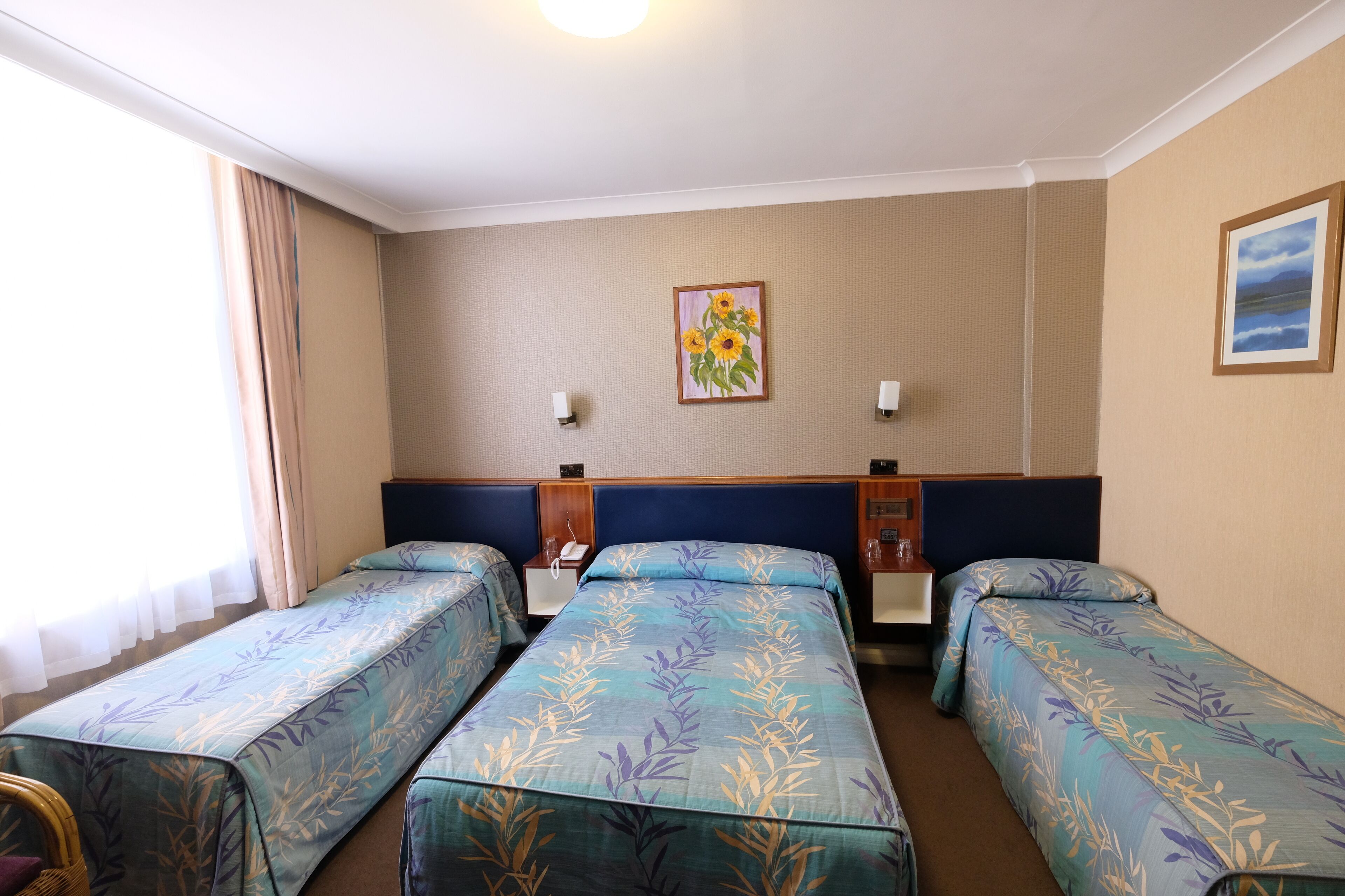 A hotel room with two single beds, a headboard, a nightstand, a lamp, and a painting on the wall.