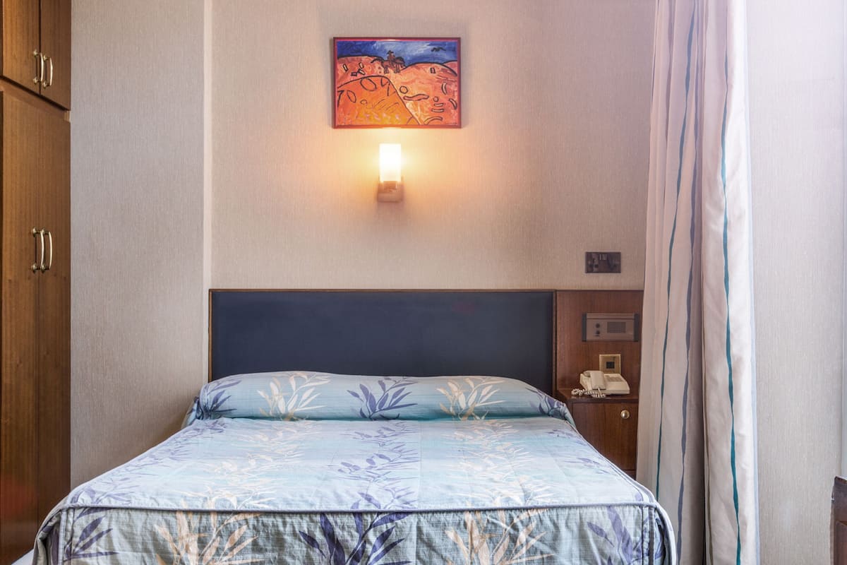 A bedroom with a bed, a wall-mounted light, a painting, and a telephone on a nightstand.