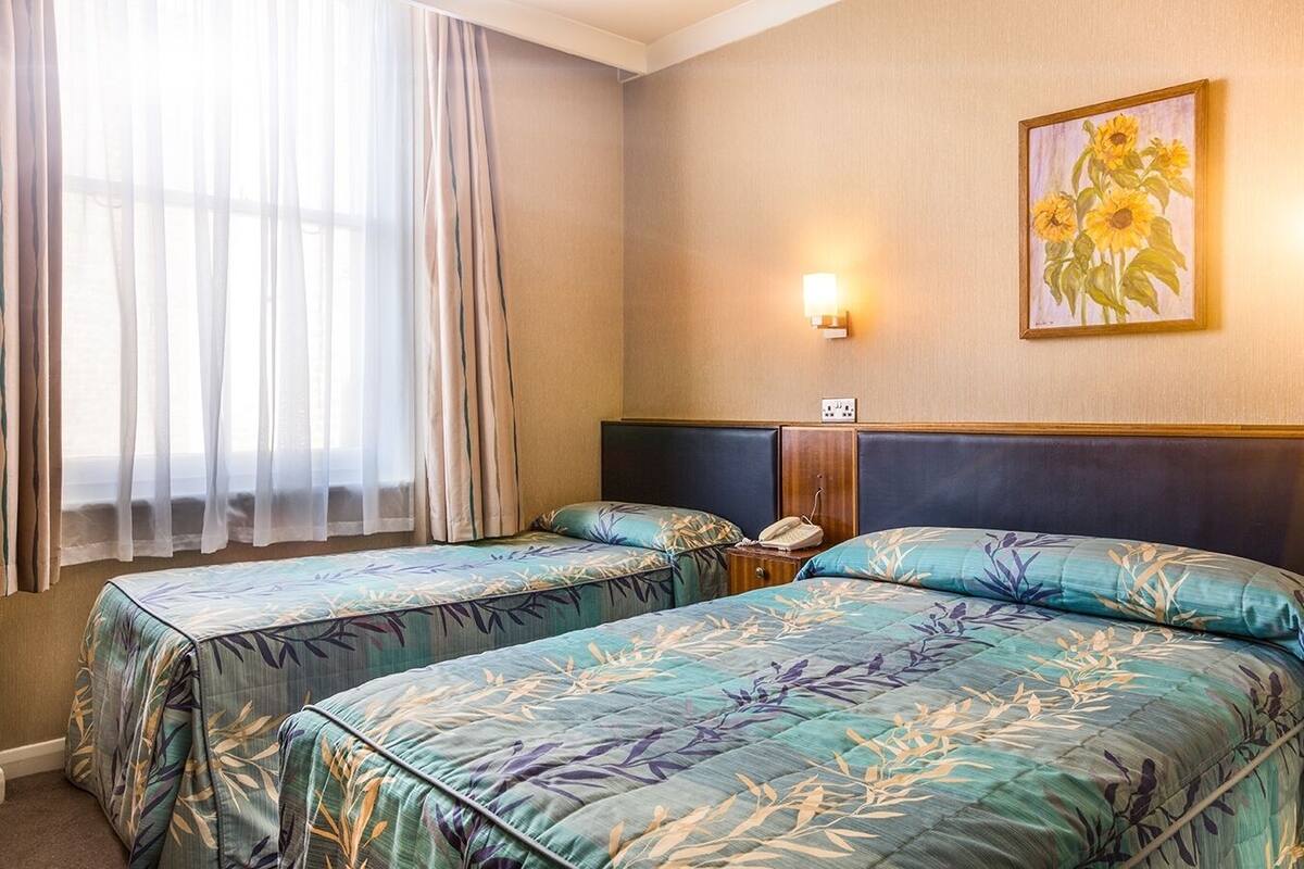 A hotel room with two beds, a nightstand, a lamp, and a painting of sunflowers on the wall.