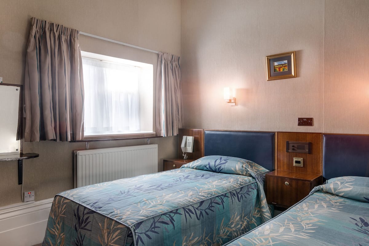 A hotel room with two beds, a window with curtains, a bedside table with a lamp, and a wall-mounted light fixture.