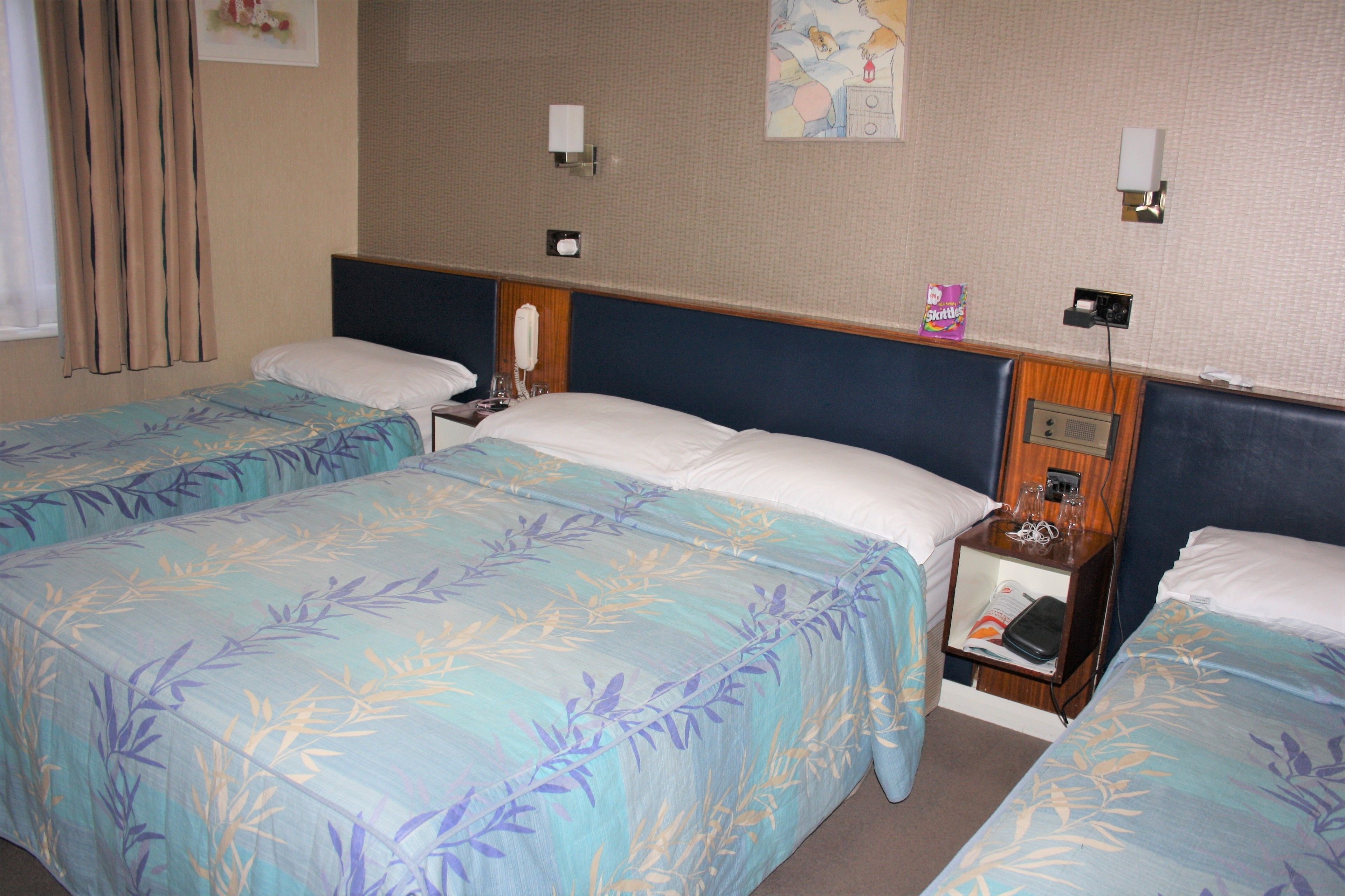 Habitación de hotel con dos camas individuales, una mesita de noche con lámpara, una pequeña mesa con un aperitivo y un teléfono en la pared.