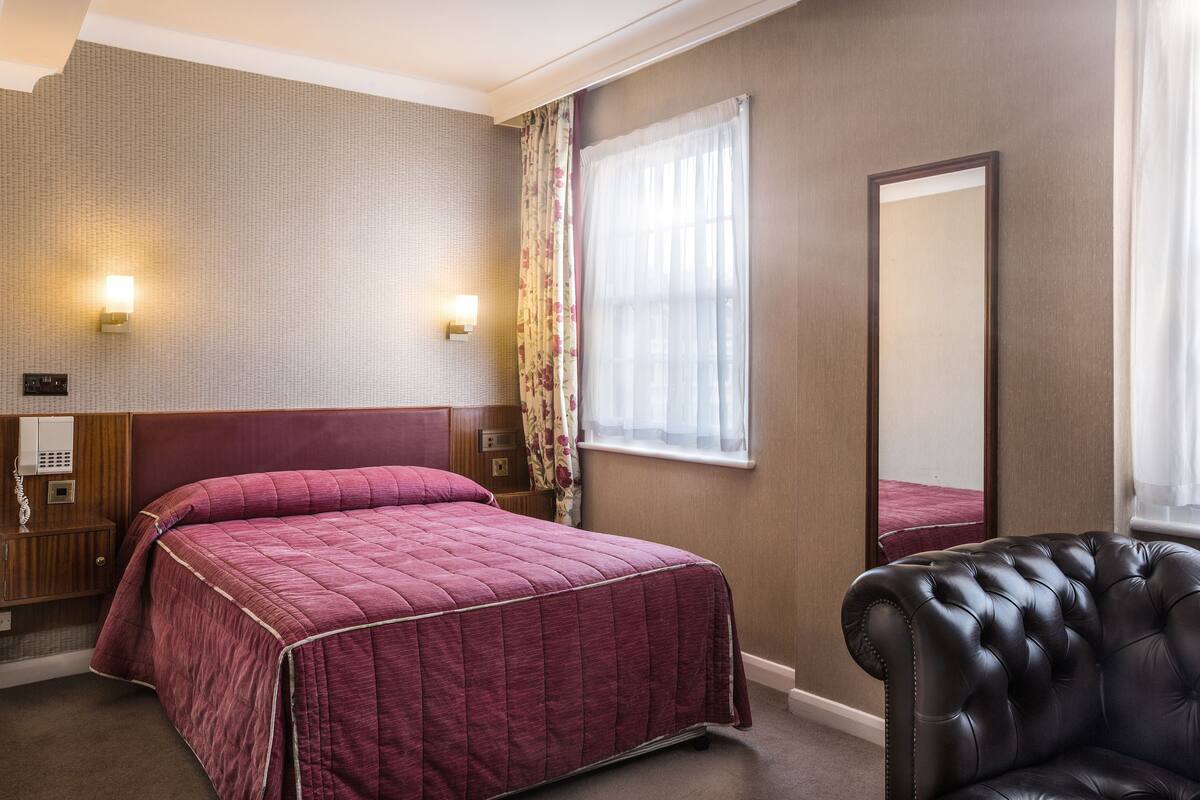 A hotel room with a large bed, a chair, a bedside table, a lamp, and a mirror.