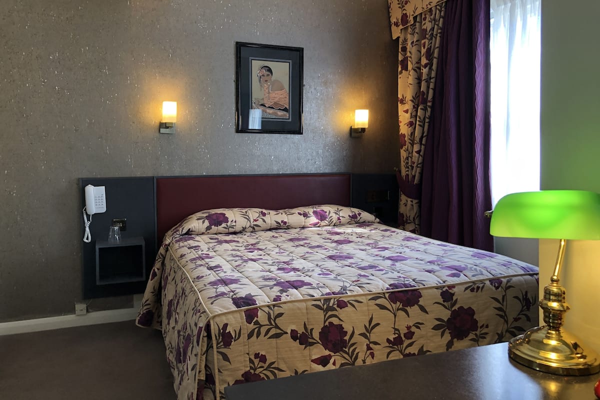 A hotel room with a bed, a bedside phone, a framed picture on the wall, and a lamp.