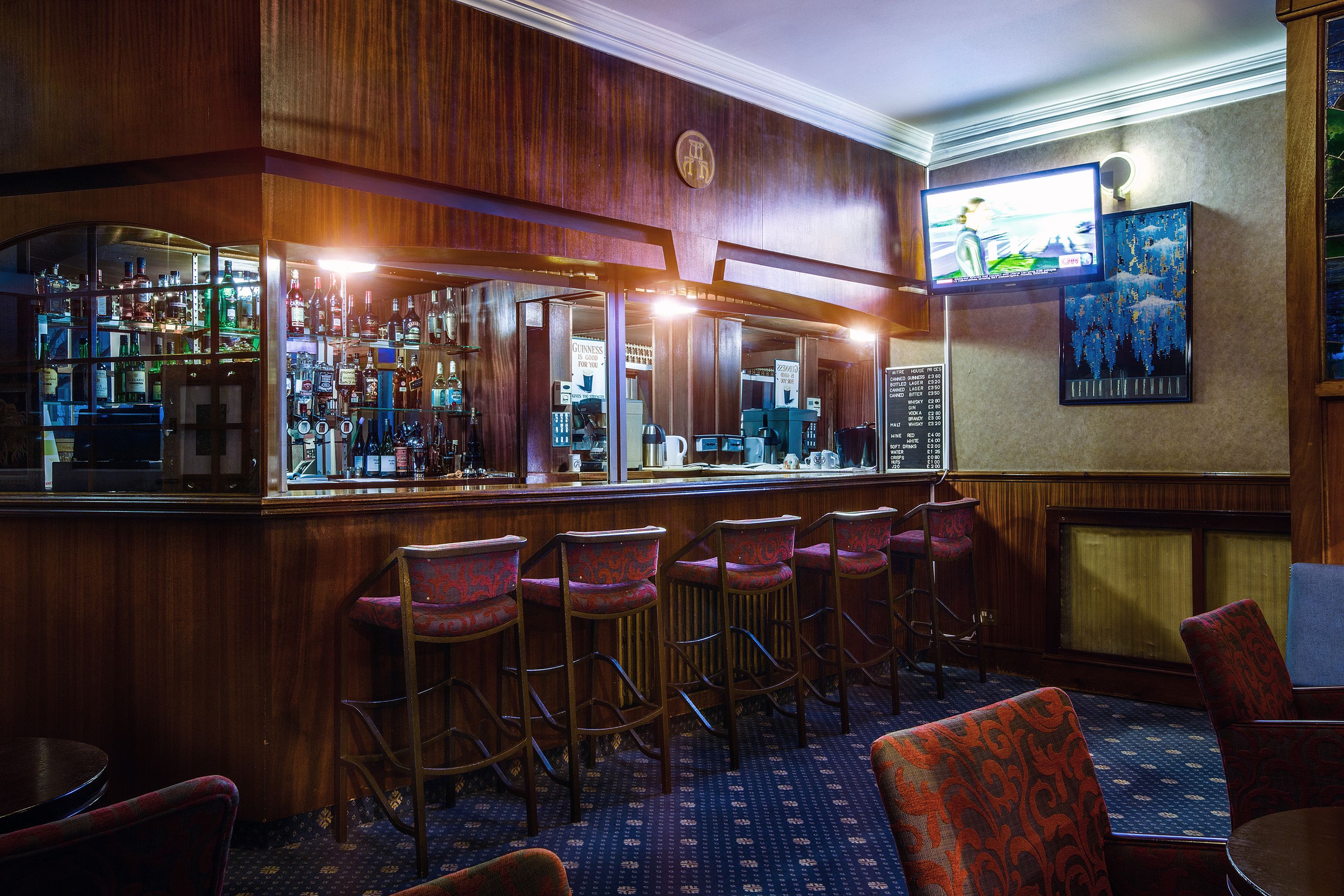 A bar area with wooden paneling, bar stools, and a counter displaying various bottles.