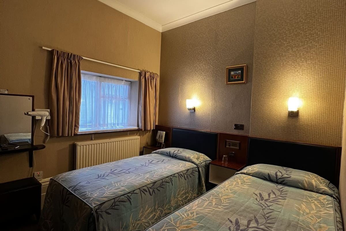 A hotel room with two single beds, a window with curtains, a radiator, a bedside table with a lamp, and a wall-mounted light fixture.