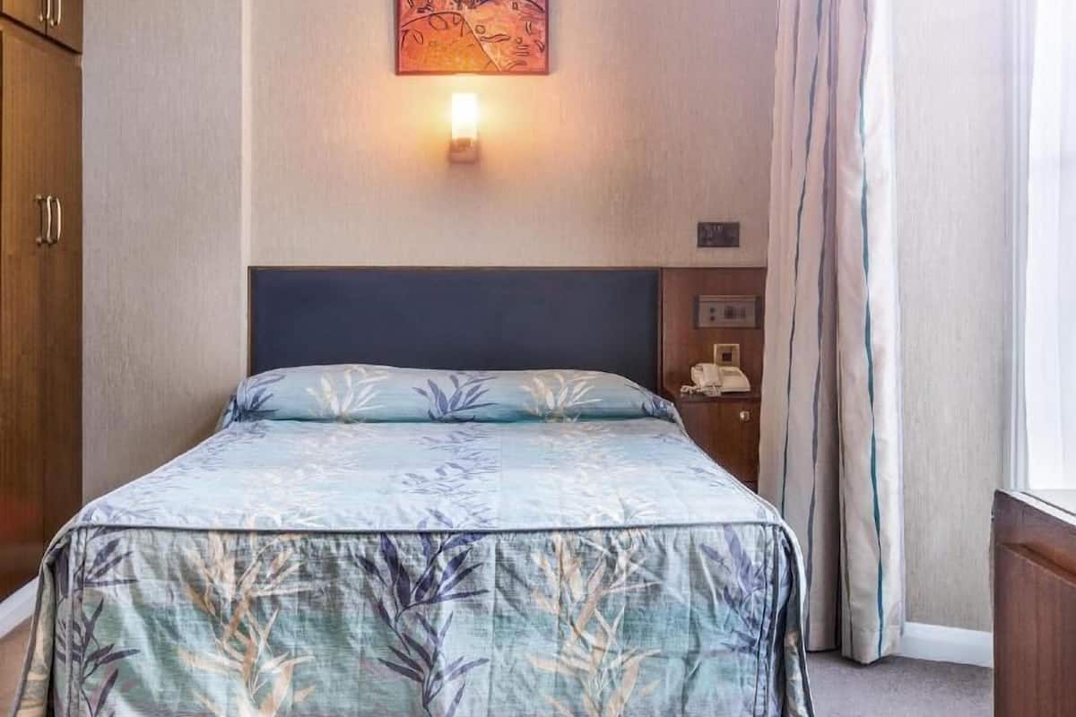 A hotel room with a bed, a nightstand, a wall-mounted light, a painting, and a window with curtains.