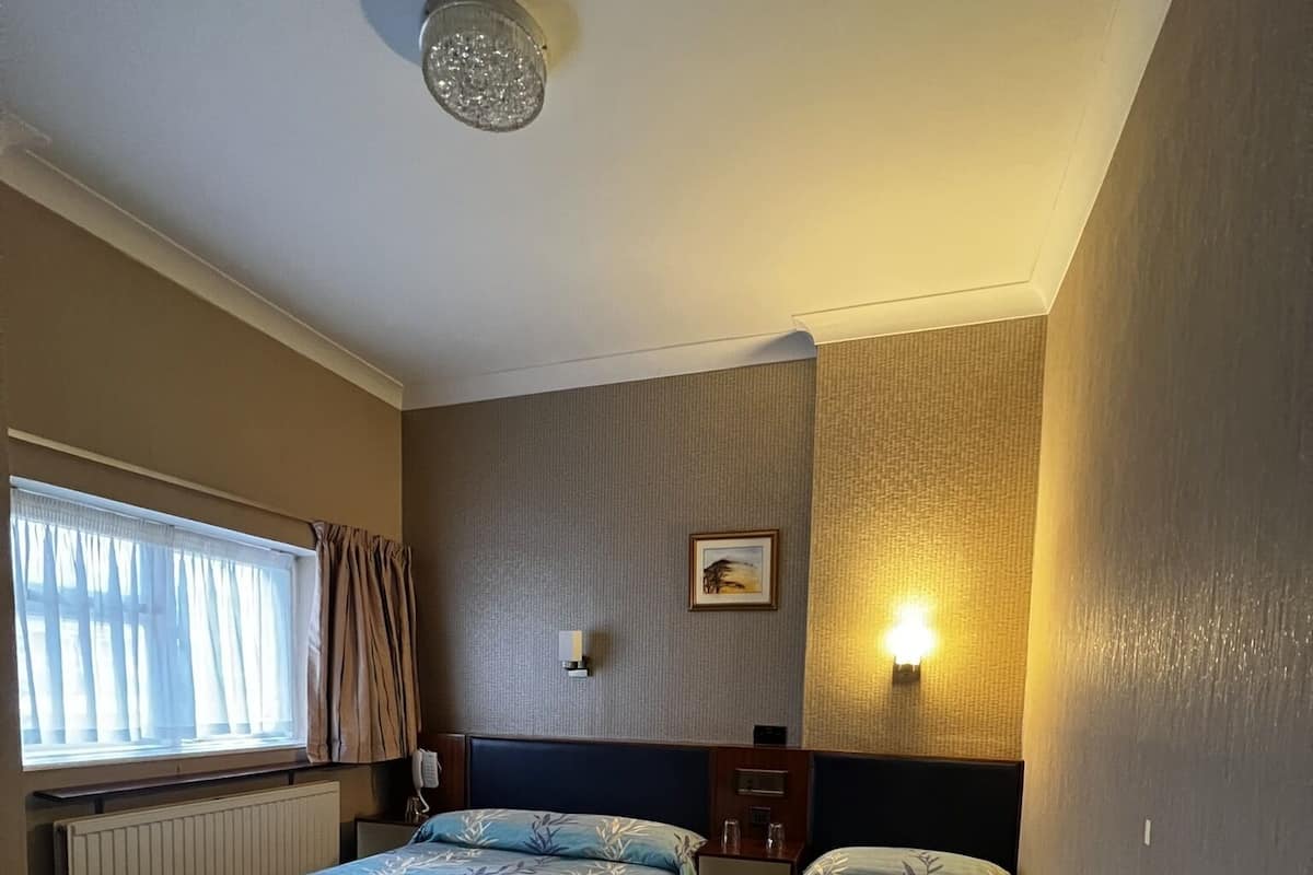 A hotel room with two beds, a window with curtains, a wall-mounted light, and a ceiling light fixture.