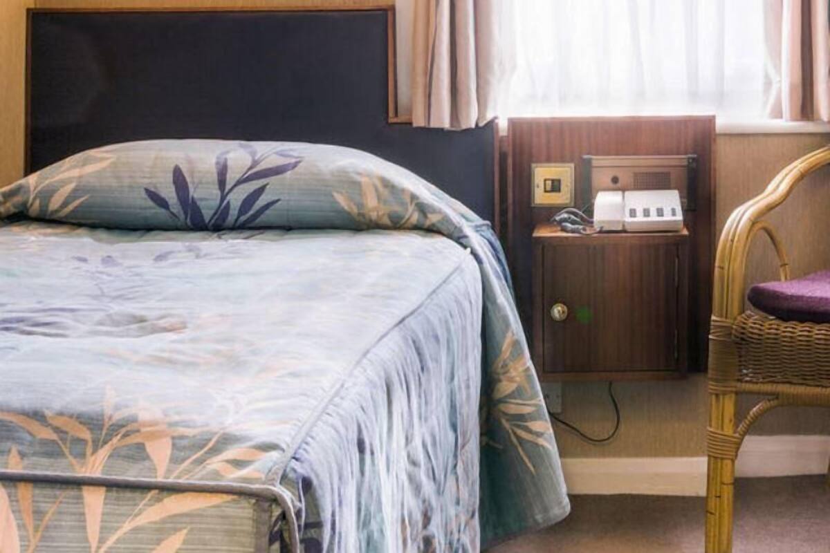 A hotel room with a bed, a nightstand with a phone and a lamp, a wicker chair, and a window with curtains.