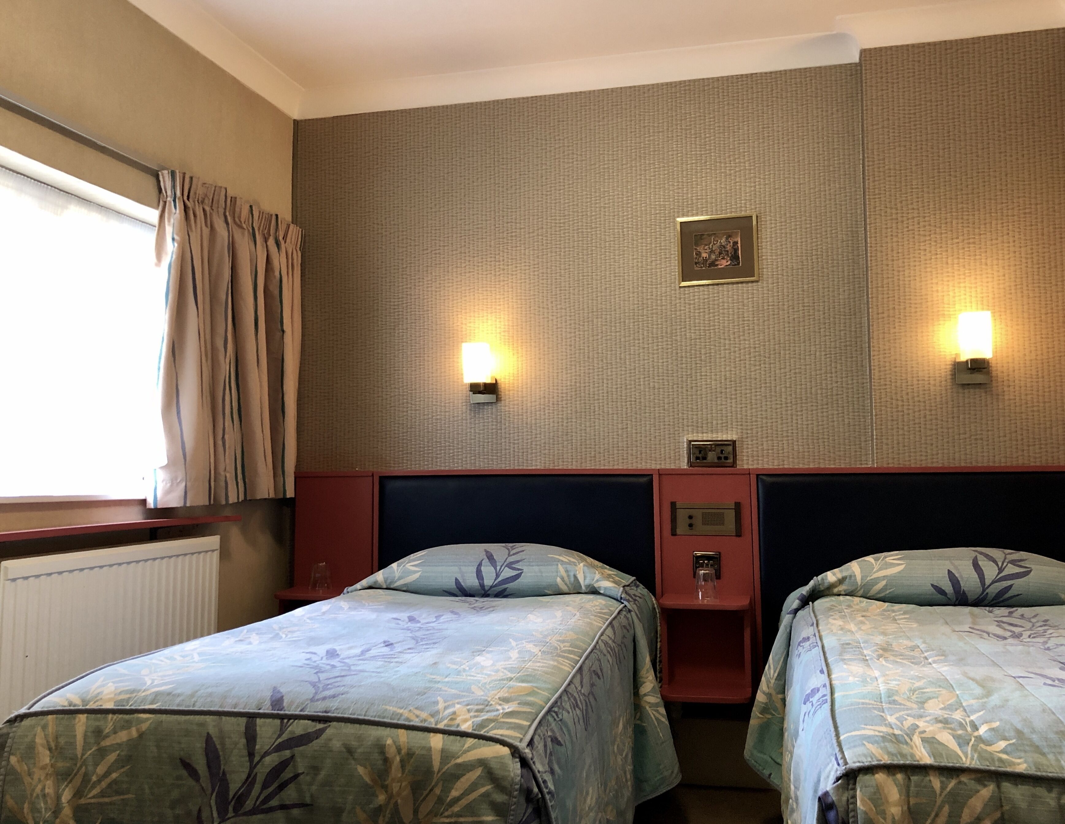 Habitación de hotel con dos camas, una ventana con cortinas, una lámpara de pared y un pequeño marco de fotos.