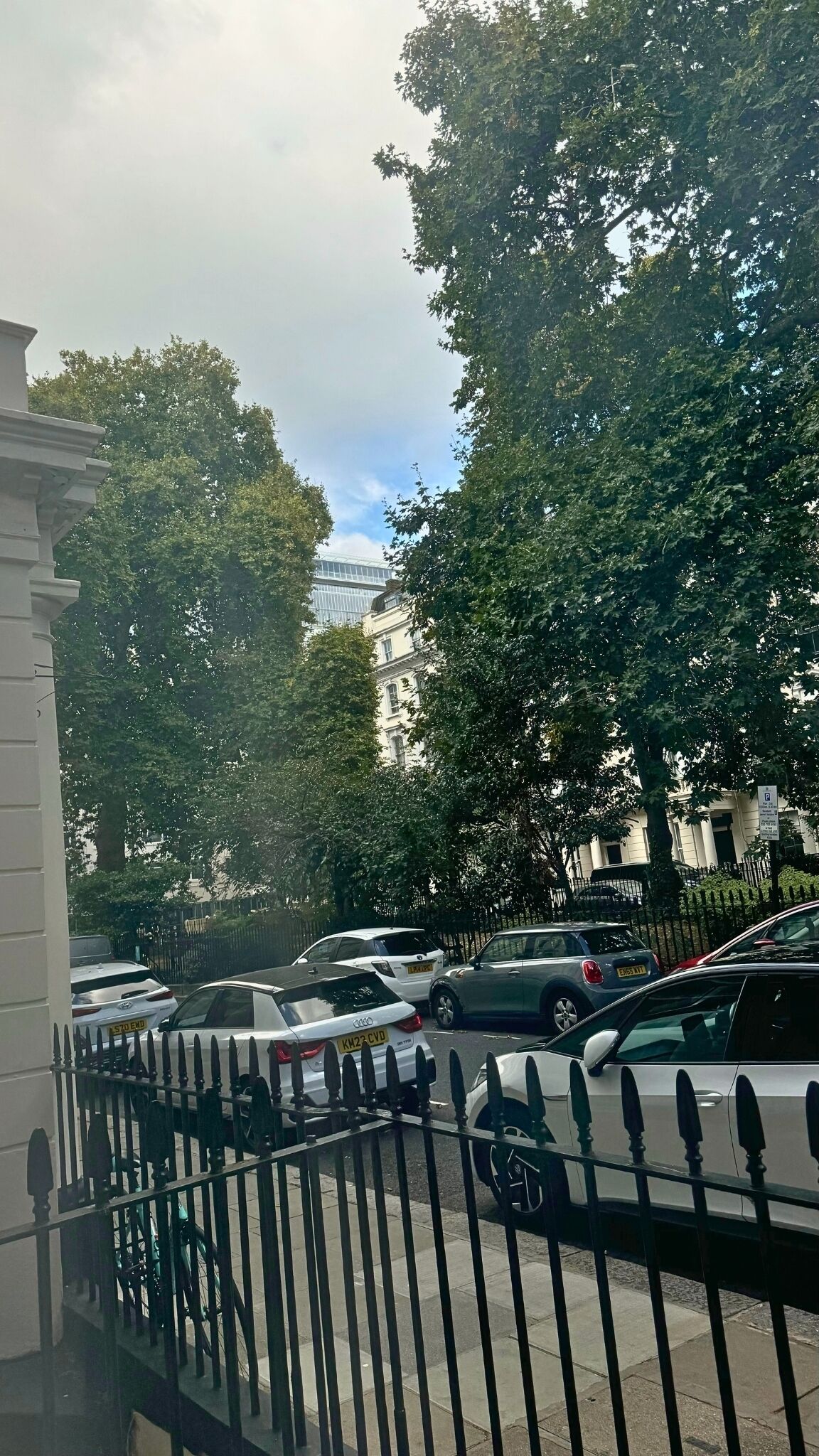 A parking area with multiple cars, a white building with columns, and trees in the background.