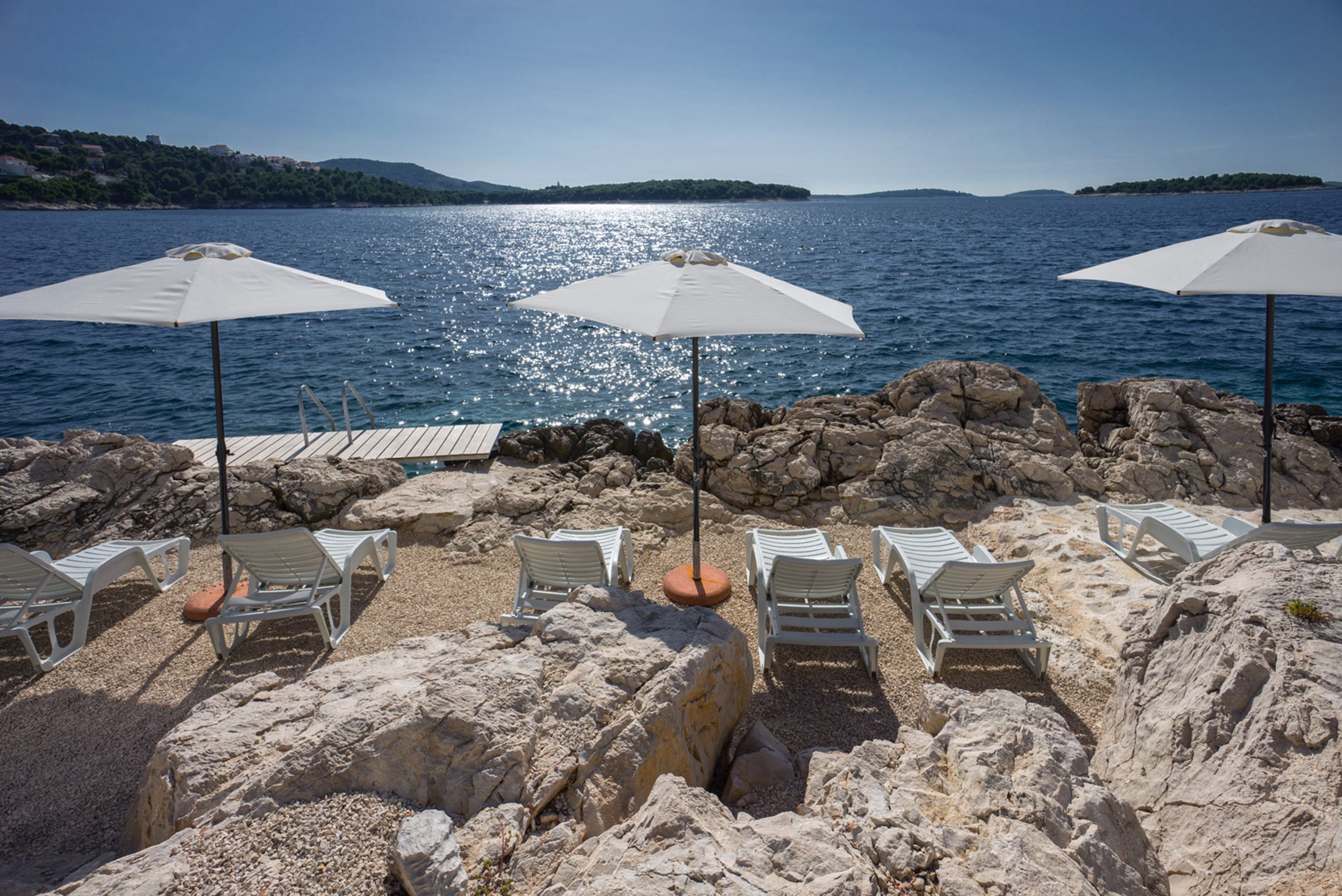 A rocky beach with white lounge chairs and umbrellas, overlooking a calm sea with a distant island.