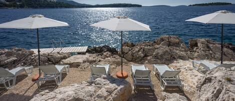Une plage de galets avec des chaises longues blanches et des parasols, donnant sur une mer calme et une île lointaine.