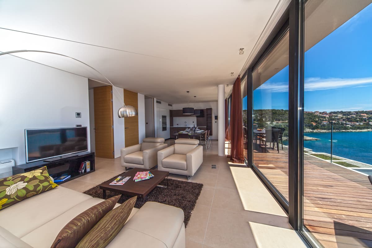 A modern living room with a flat-screen TV, sofa, and coffee table. There is a dining area with a table and chairs. A balcony with a view of the sea is visible.
