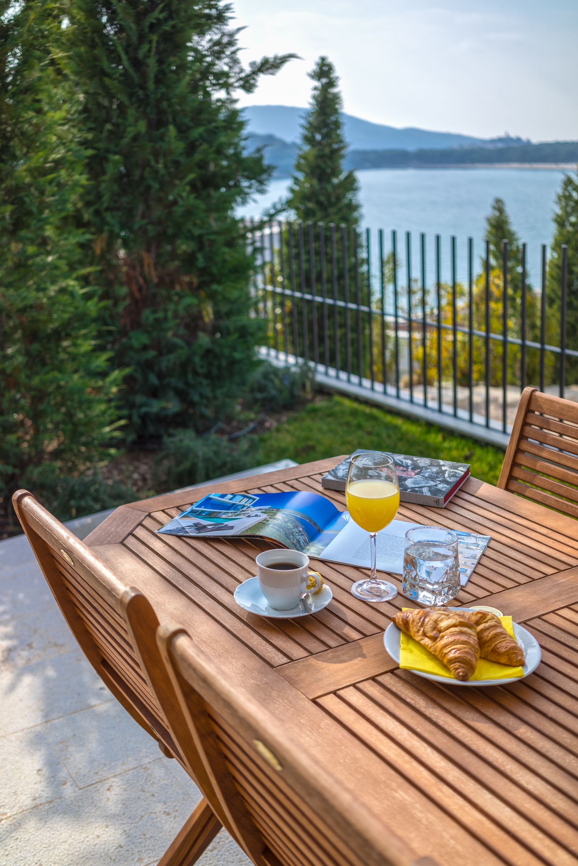 Ein Holztisch mit einem Croissant, einem Saftglas, einer Tasse und einer Zeitschrift auf einem Balkon mit Blick auf einen See.