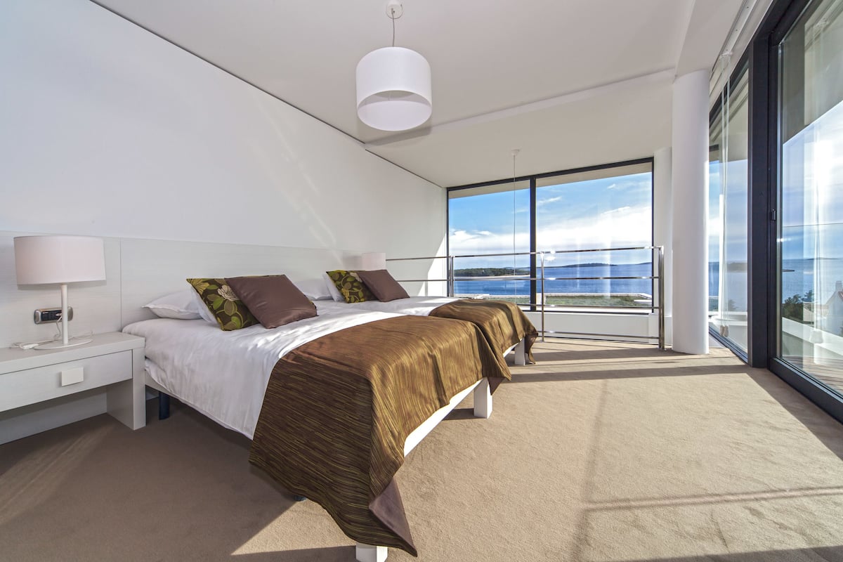 A modern bedroom with two beds, a large window with a view, a white nightstand, and a hanging light fixture.
