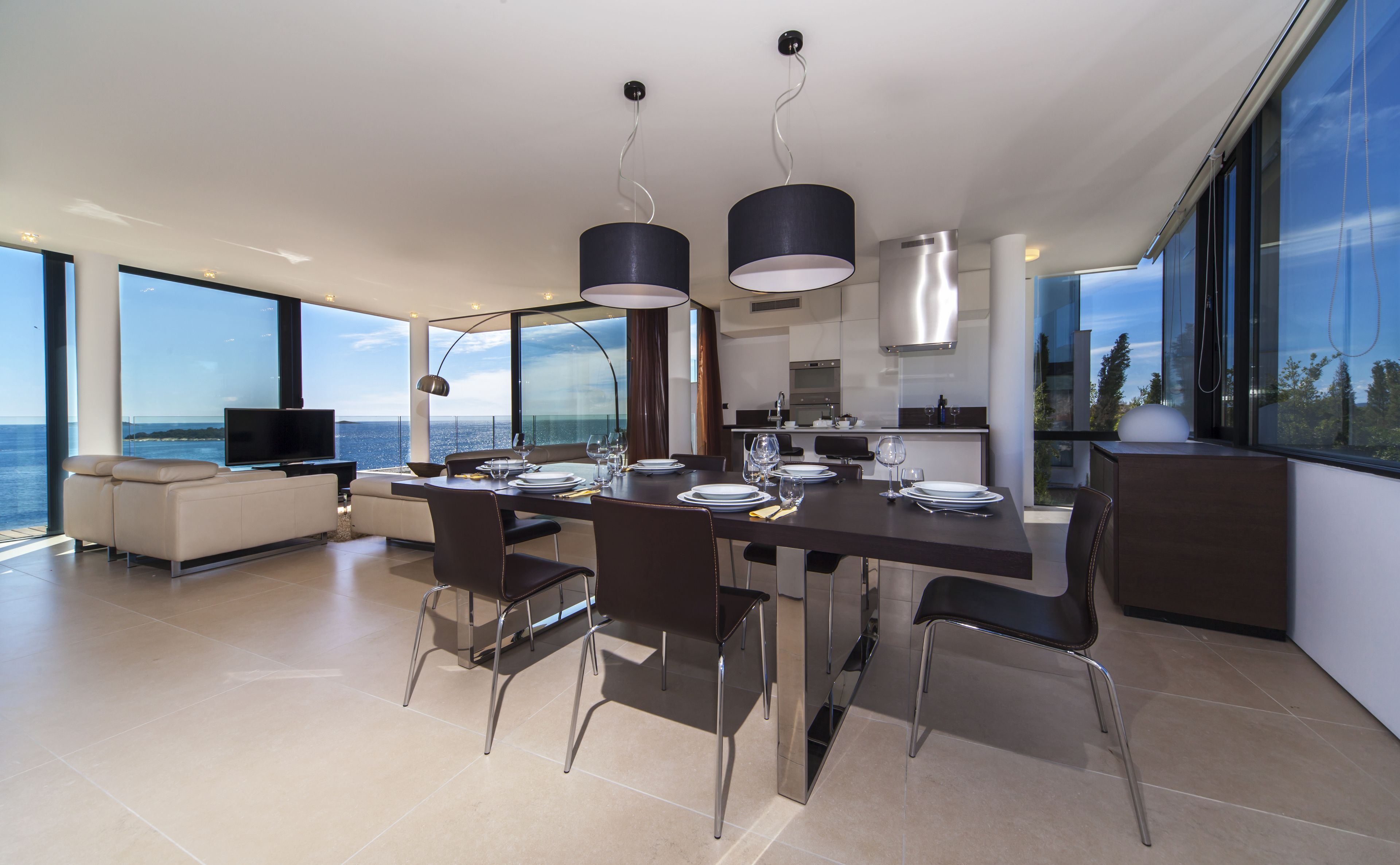 A modern dining area with a large table, chairs, and pendant lights. There is a television in the background and a view of the sea through large windows.