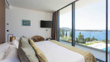 A hotel room with a large bed, a flat-screen TV, a chair, and a view of the sea.