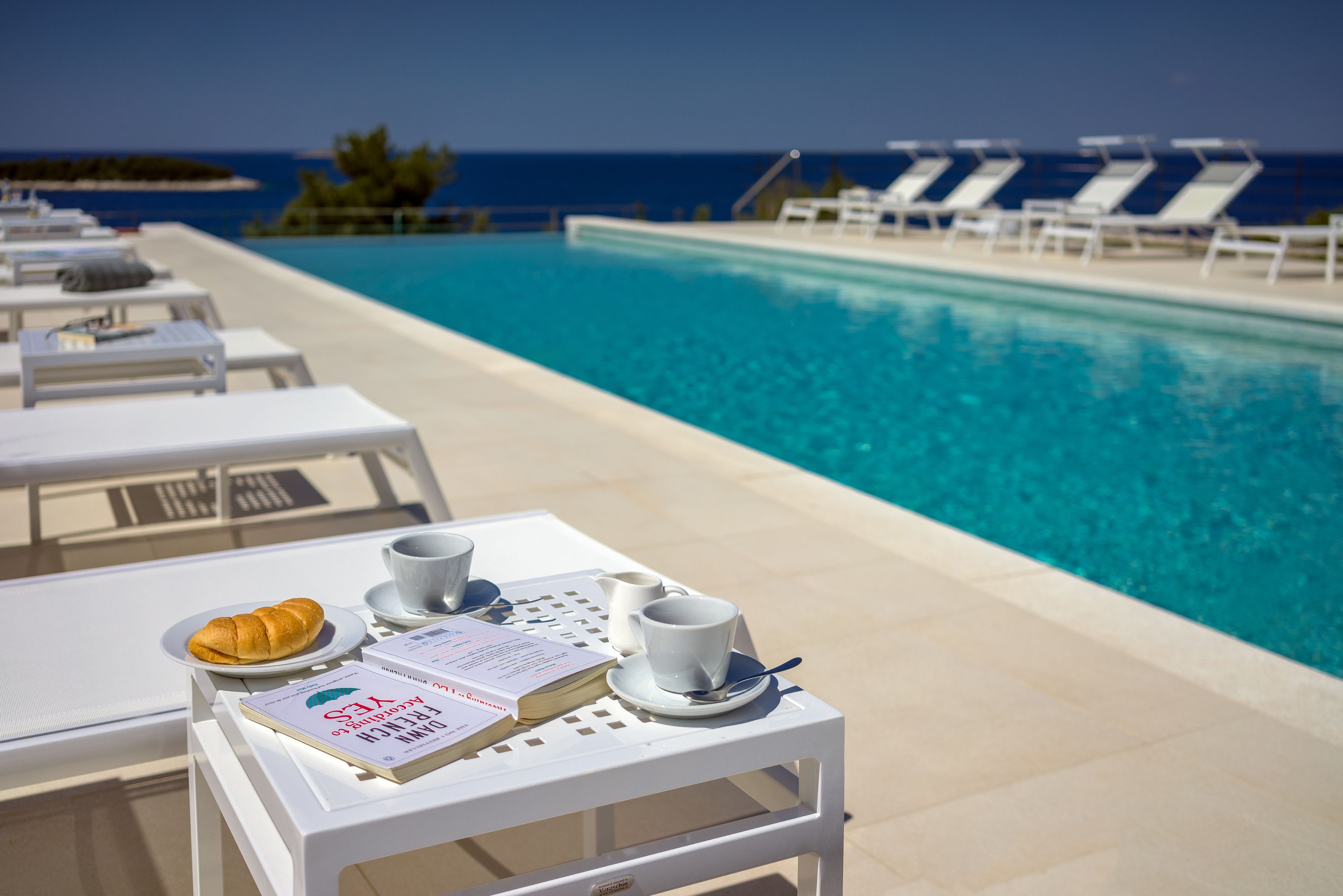 A poolside table with a croissant, a cup, and a menu, overlooking a pool with lounge chairs and a view of the ocean.
