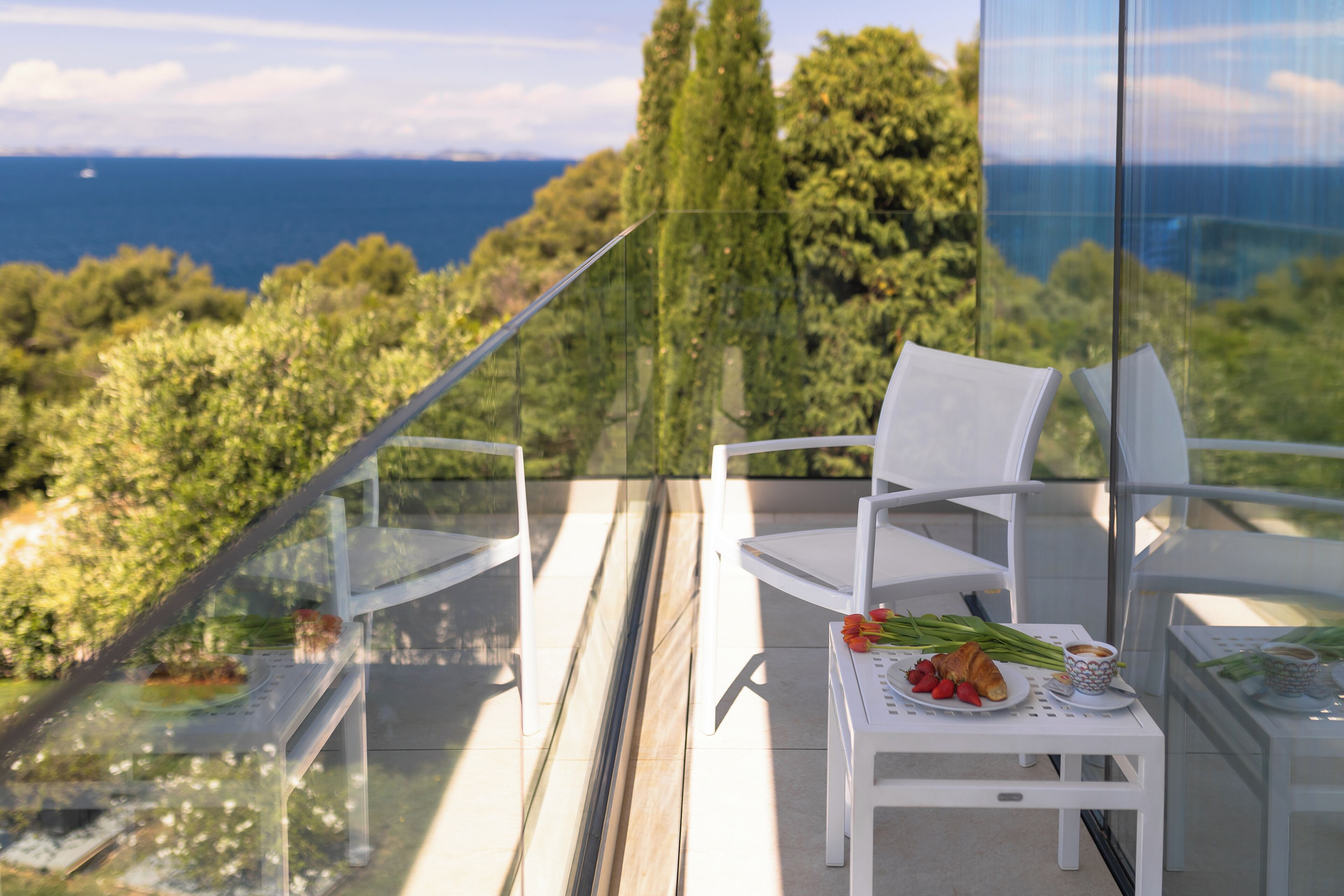 A balcony with a glass railing, white chairs, a small table with a plate of food, and a view of the sea and trees.
